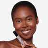 Smiling person holding a clear lip gloss tube with a black cap, showcasing radiant skin and glossy lips.