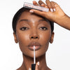 Model holding with one hand a clear/pink cosmetic tube to her forehead while applying product to her lips with the other, showcasing a glossy makeup look against a plain background