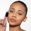 Model showcasing a glossy nude lip color, holding a clear tube with a black cap labeled "OFIR", against a plain background.