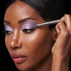 A person applies shimmery lavender eyeshadow with a makeup brush, highlighting their eyelid.