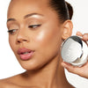 A model showcasing smooth, glowing skin holds a sleek, round compact with a reflective silver finish and branded text, emphasizing a polished and radiant complexion.