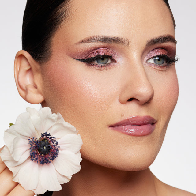 Elegant woman with smooth, glowing skin, wearing subtle pink and purple eye makeup and glossy lips, holding a delicate white flower near her face.