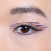 Close-up of an eye with purple eyeliner applied in bold, parallel strokes extending outward from the outer corner, complemented by defining mascara on the eyelashes.