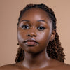 A close-up of a woman with glowing skin and styled braids, showcasing natural beauty against a neutral background.
