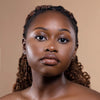 A woman with smooth, glowing skin and styled braids poses against a neutral beige background, highlighting her natural beauty and glossy lips.