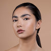 A model showcases a natural makeup look with glowing skin, subtle eye makeup, and a nude lip against a neutral background.