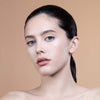 A close-up of a woman with smooth skin, subtle makeup, and blue eyes, against a soft beige background.
