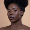 A woman with radiant, smooth skin and glossy dark lip color is photographed against a soft beige background, highlighting the makeup's finish and complexion-enhancing effect.