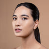 A model with flawless, radiant skin and subtle makeup, featuring defined brows and glossy lips, set against a soft beige background.