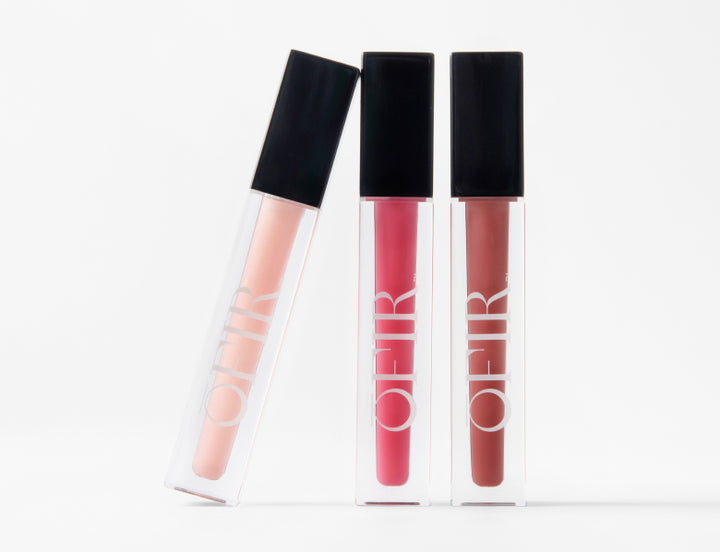 Three Ofir lip glosses in nude, pink, and mauve shades displayed upright on a white background