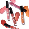 Flat lay of lip gloss tubes in assorted pink, mauve, coral, and golden shades, arranged on white background with swatches showing glossy finishes.