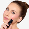 Model holding a deep pink blush stick in a black tube, showcasing a light pink blush pigmentation in a radiant natural makeup look.