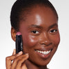 Model holding near her cheek  a purplish blush stick in a black tube, showcasing a purplish blush pigmentation in a radiant natural makeup look.