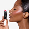 Model holding and looking sideways at a deep pink blush stick in a black tube, showcasing a pink blush pigmentation in a radiant natural makeup look.