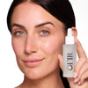 A woman with clear, radiant skin holds a frosted glass bottle of serum with a dropper cap, labeled "OFIR," highlighting its skincare benefits.