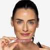 Model holding a dusty rose pink lip gloss tube in one hand, with the shade applied to her lips, showcasing a glossy pink makeup look
