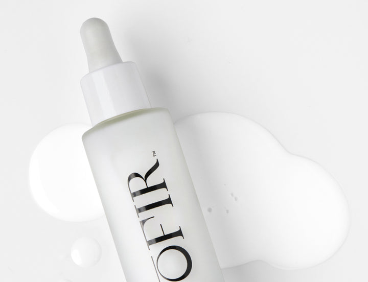 A frosted white OFIR Face Milk bottle with a dropper top, lying on a surface with white liquid droplets and a spill pooled around it.