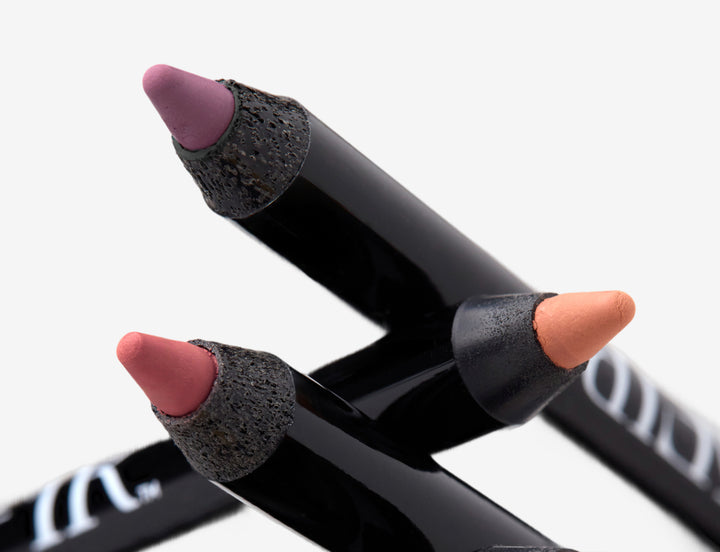 Three eyeliner pencils in shades of purple, pink, and peach, with sleek black casings arranged on a neutral background.
