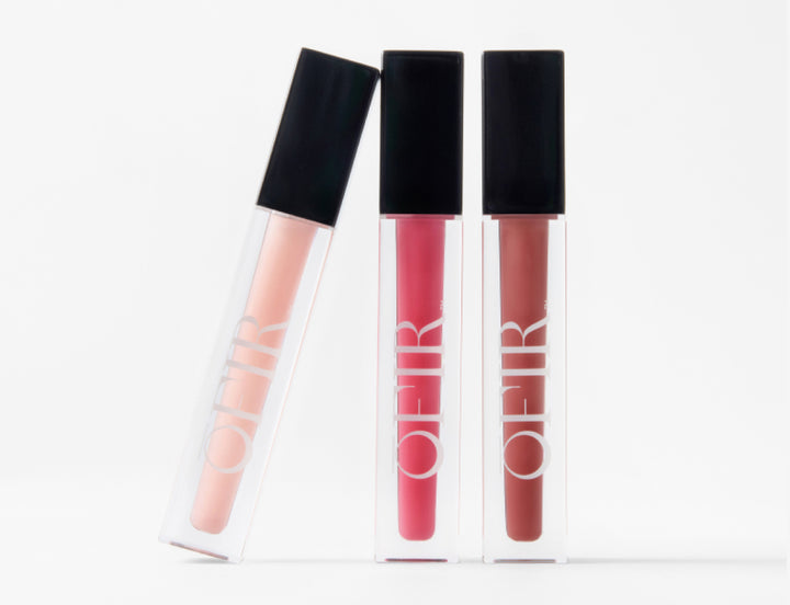Three liquid lipsticks in clear tubes with black caps, featuring shades of pale pink, vibrant red, and warm brown, labeled "OFIR".