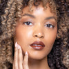 Woman showcasing glossy brown lipstick and subtle eye makeup, with defined curls framing her face, and a hand gently touching her cheek with nude manicured nails.