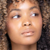 Woman with glowing skin and glossy nude lips, featuring natural curly hair and subtle makeup.