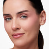 Woman with subtle makeup, featuring soft pink eyeshadow and blush, minimal mascara, and glossy nude lips, with dark hair pulled back.