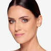 Woman with smooth, radiant skin and glossy lips, showcasing elegant makeup featuring smoky eyeshadow, long lashes, and neatly groomed eyebrows. Her dark hair is styled back, accentuating her refined and polished appearance.