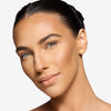 Person with smooth, glowing skin and subtle makeup, showcasing clear complexion and natural lip color. Hair styled in a sleek ponytail against a plain background.