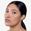 Person with smooth, glowing skin and subtle makeup, showcasing clear complexion and natural lip color. Hair styled in a sleek ponytail against a plain background.