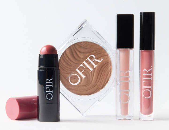 A group of OFIR makeup products displayed together, including a berry-toned cream blush stick with its cap off, a pressed bronzer in a clear compact, and two lip glosses in pink and nude shades with black caps.