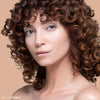A woman with curly brown hair and natural makeup poses against a neutral background, showcasing flawless skin.