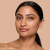 A person with smooth, glowing skin and natural makeup, wearing a small gold nose ring against a neutral background, showcasing a beauty product.
