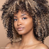 A person with beautifully styled curly hair and natural makeup looks confidently at the camera against a neutral background, showcasing a radiant complexion.