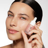 A person with smooth, glowing skin holds a pink serum bottle with a dropper up to their face, showcasing the product.
