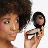 A person holding a compact mirror with a soft pink blush applied, showcasing smooth, radiant skin and a subtle makeup look.