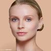 Close-up of a person with smooth, natural makeup and a serene expression against a neutral background, promoting the makeup shade "Bare Rice."