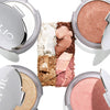 Four open makeup compacts surround a central arrangement of crushed highlighters and blush powders in shimmering shades of white, bronze, pink, and peach.