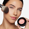 Woman applying blush with a brush while holding a compact labeled "OFIR," showcasing smooth and natural makeup application.
