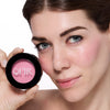 Woman with light skin tone holding a pink blush compact labeled "OFIR" against her cheek, highlighting natural makeup application.
