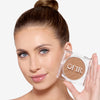 Model holding a compact bronzer while showcasing a light nude shade on her cheekbones and a flawless makeup finish. 