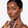 Model holding a compact bronzer while showcasing a dark nude shade on her cheekbones and a flawless makeup finish.