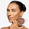 Model holding a compact bronzer while showcasing a medium nude shade on her cheekbones and a flawless makeup finish.