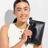 Smiling individual holding a black, drawstring makeup bag with embossed branding, dressed in a sleeveless white top with button details.