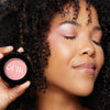 Radiant complexion enhanced with soft pink blush, held beside a smiling face with closed eyes, showcasing smooth curly hair.
