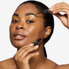 Model showcasing a natural but enhance black brow design and color in a radiant natural makeup look while holding the serum next to her face and about to apply to her brows