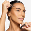 Model showcasing a natural but enhance brown brow design and color in a radiant natural makeup look while holding the serum next to her face and about to apply to her brows