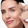 Smiling person holding an open compact highlighter near their face, showcasing smooth, luminous skin with a subtle glow.