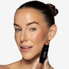 Model holding near her cheek  a pink/light brown-ish blush stick in a black tube, showcasing a purplish blush pigmentation in a radiant natural look