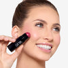 Model holding near her cheek  a depp magenat blush stick in a black tube, showcasing a purplish blush pigmentation in a radiant natural look