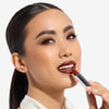 Woman applying deep red lipstick, highlighting smooth skin and subtle eye makeup, against a plain background.